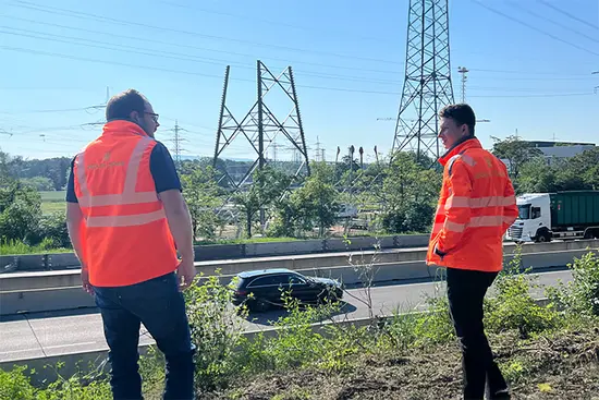 Zwei Personen in orangefarbenen Sicherheitswesten stehen an einer Straße mit Stromleitungen und Autos im Hintergrund.