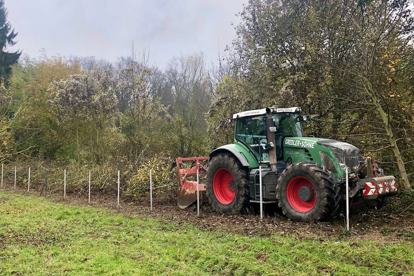 Ein grüner Traktor mit roten Rädern pflügt den Boden neben neuen Zaunpfählen – Baum- und Grünpflege für gesunde Bäume in der Nähe des Waldes.