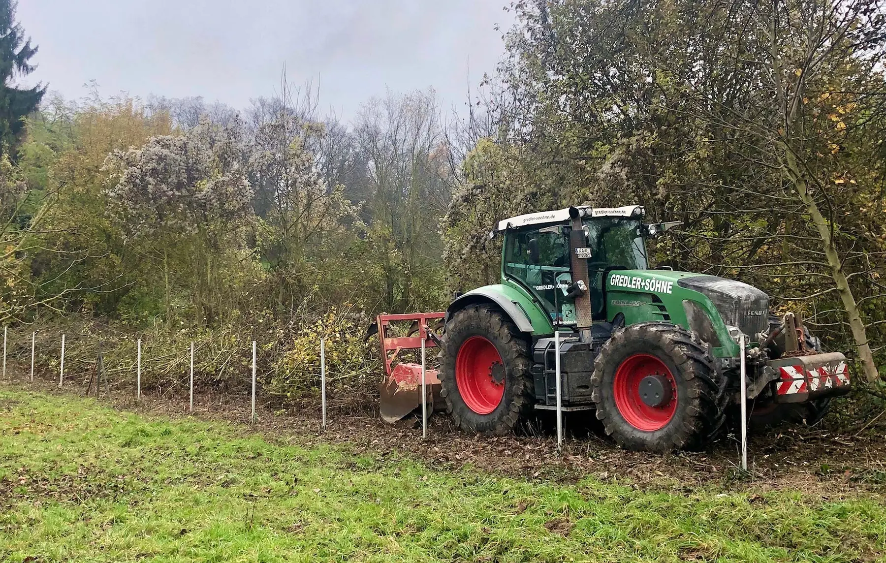 Ein grüner Traktor mit roten Rädern fährt an einem Zaun entlang, der neben dichten Bäumen auf einer Wiese liegt.