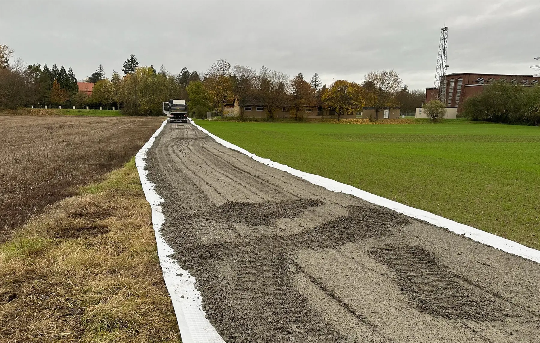 An einem bewölkten Tag verläuft zwischen einem Feld und einer mit weißen Streifen gesäumten Rasenfläche ein im Bau befindlicher Schotterweg.