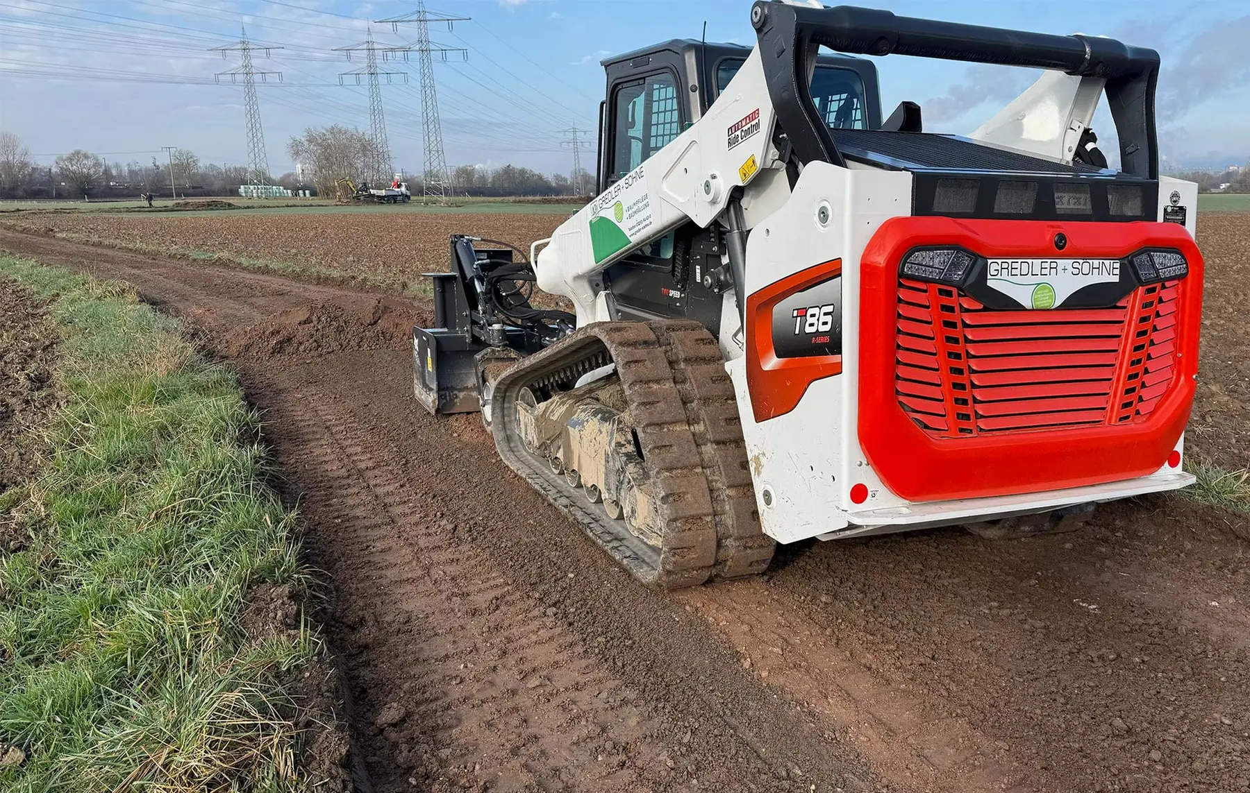 Ein rot-weißer Raupenlader ebnet einen Feldweg in einer ländlichen Gegend in der Nähe von Stromleitungen und Feldern.