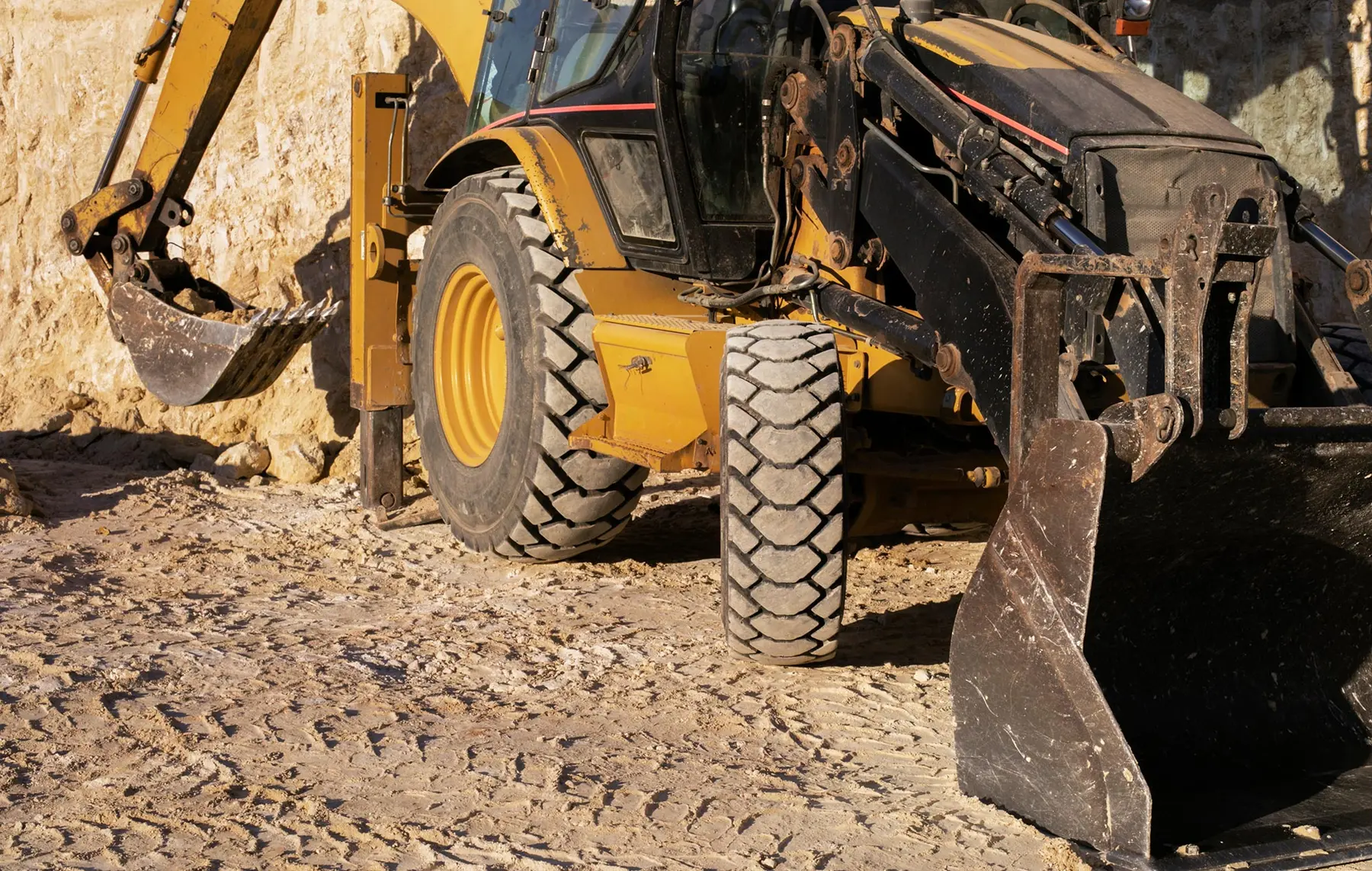 Gelber Baggerlader mit großen Reifen und zwei Baggerschaufeln, geparkt auf einer unbefestigten Baustelle.
