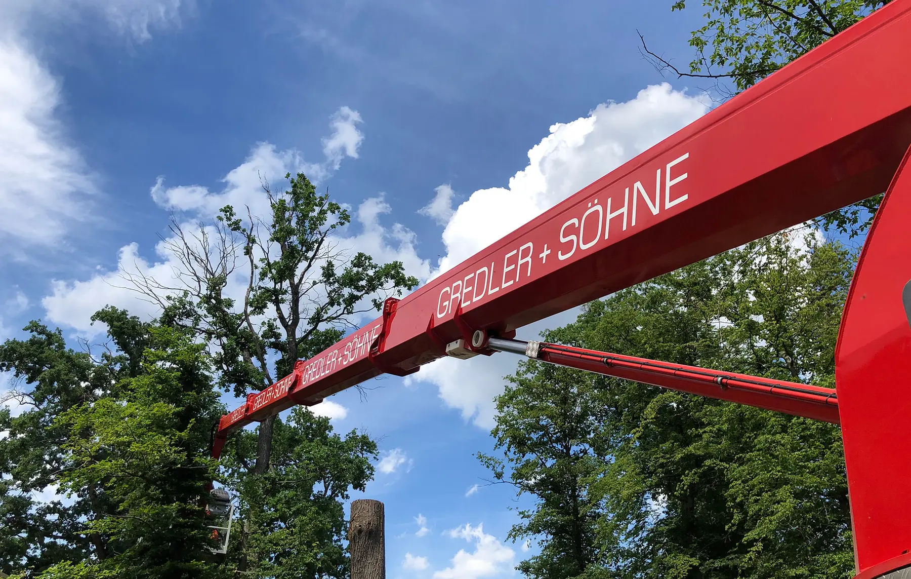Roter Kranarm mit dem Text GROßDLER + SÖHNE ausgefahren für Baumfällung neben grünen Bäumen unter einem blauen Himmel mit weißen Wolken.