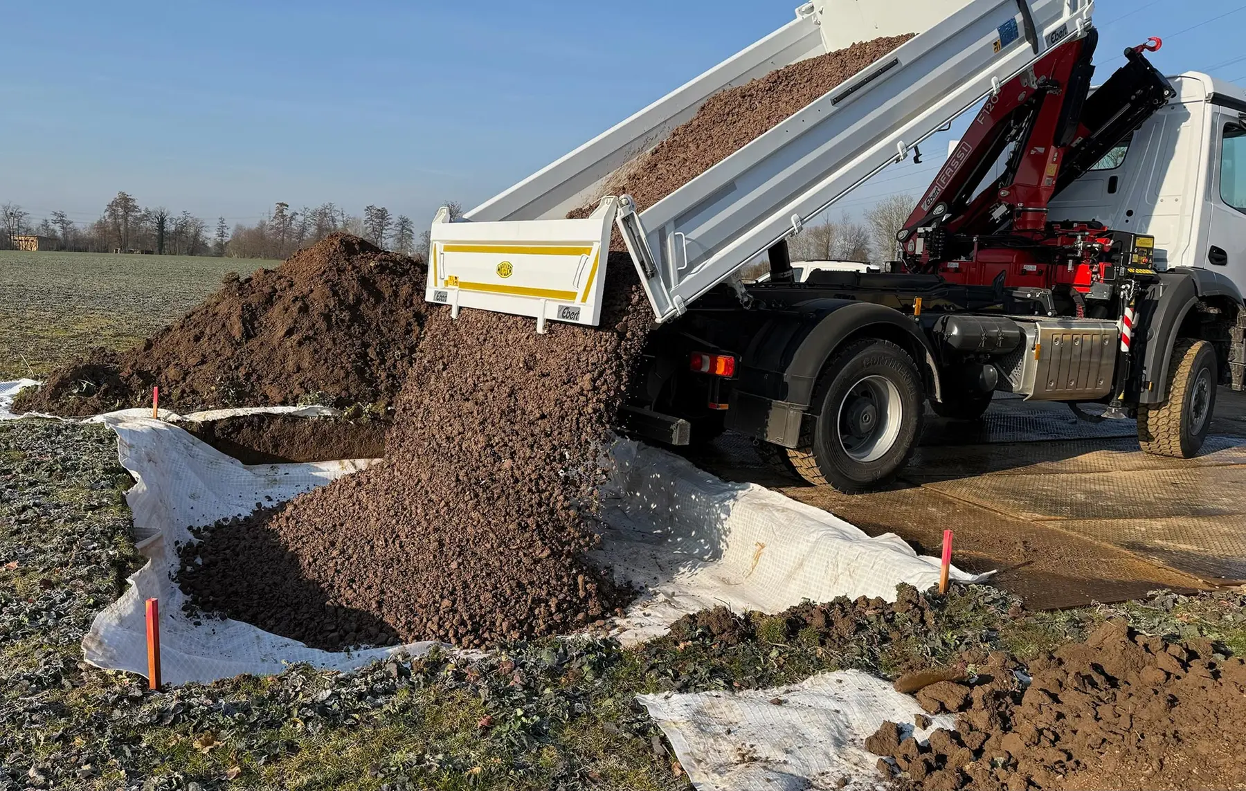 Ein Muldenkipper lädt einen Haufen Kies auf einer vorbereiteten Baustelle ab, die mit einer weißen Plane abgedeckt ist.