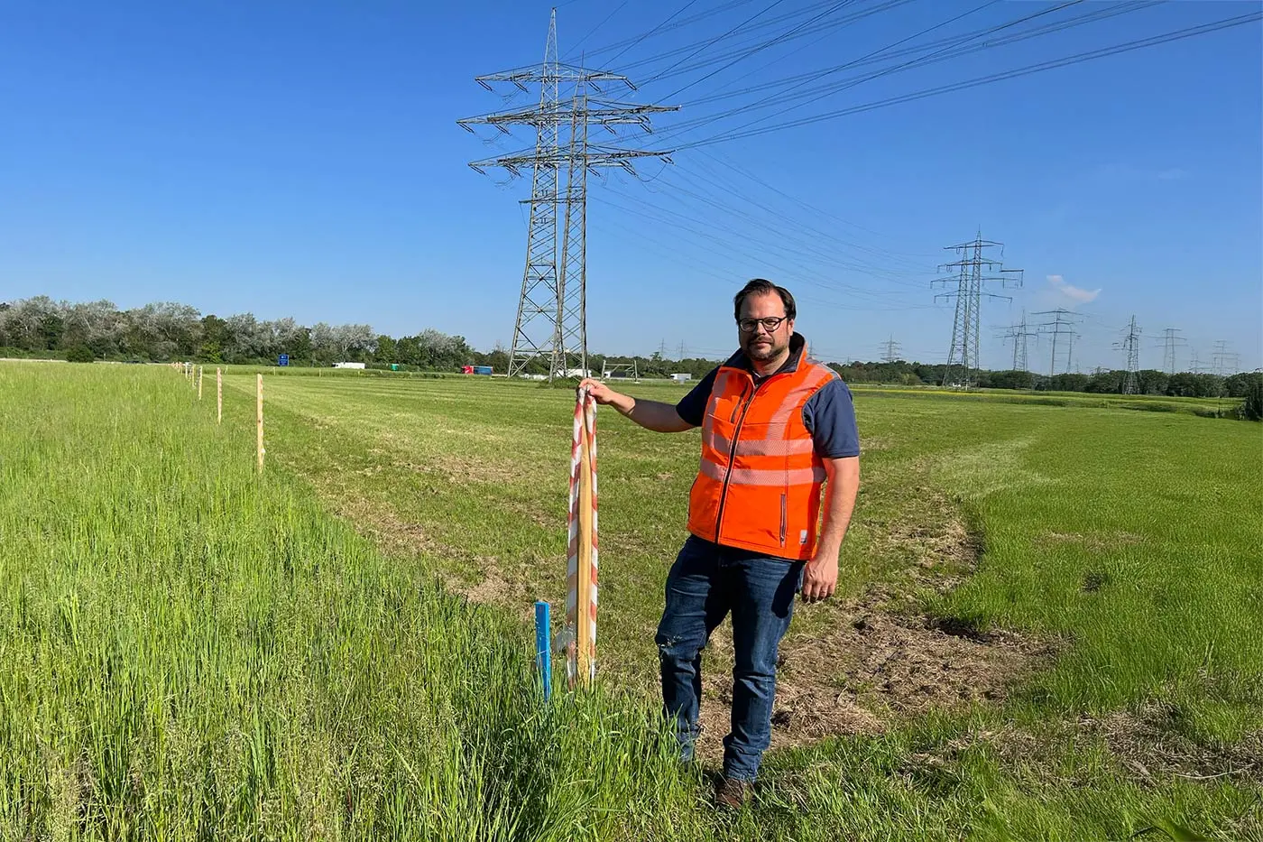 Ein Mann in einer orangefarbenen Sicherheitsweste steht auf einer Wiese neben einem Holzpfahl, im Hintergrund sind Stromleitungen und Türme zu sehen.