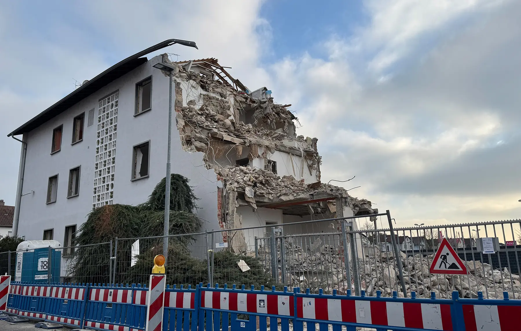 Teilweise eingestürztes weißes Gebäude hinter einem Bauzaun, Schutt und Trümmer auf dem Boden.
